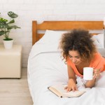 African-american girl reading book and drinking coffee in cozy bedroom, empty space