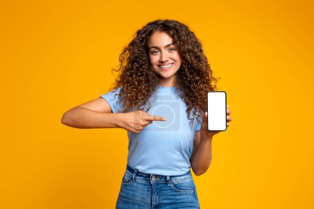 Téléchargez les photos : Joyeux geste de femme vers un écran de smartphone vierge avec un sourire, sur un fond jaune, espace de copie maquette - en image libre de droit