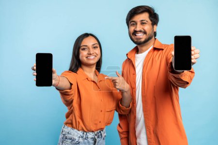 Téléchargez les photos : Couple indien souriant en chemises orange présentant un écran de téléphone vierge adapté au contenu publicitaire - en image libre de droit