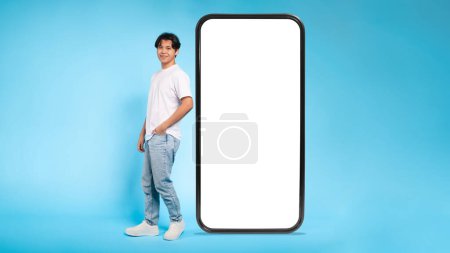 Photo for Asian guy stands casually next to a giant mockup smartphone screen with a blank display for content, blue background - Royalty Free Image