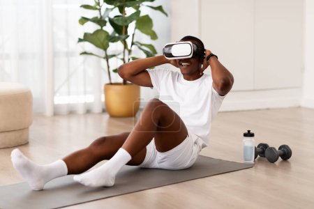 Téléchargez les photos : Un jeune noir qui exécute une séance d'entraînement au sol dans une maison moderne, en utilisant un casque VR, un exercice virtuel - en image libre de droit