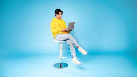 Téléchargez les photos : Adolescent asiatique garçon assis sur un tabouret haut en utilisant un ordinateur portable avec une expression contemplative sur un fond bleu uniforme - en image libre de droit