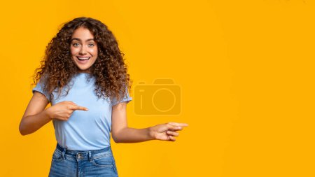 Photo for Surprised woman pointing to the side and looking at something interesting on a yellow backdrop, copy space - Royalty Free Image