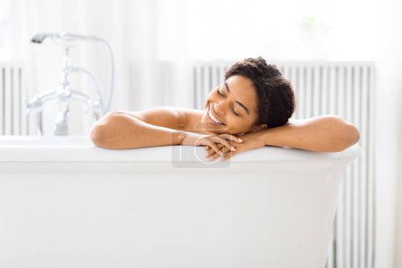 Téléchargez les photos : Femme afro-américaine détendue les yeux fermés, la tête posée sur le bord d'une baignoire, profitant d'un moment paisible - en image libre de droit