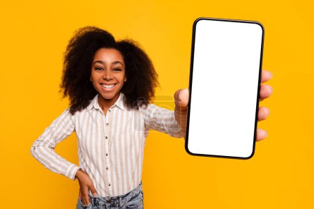 Joyeux jeune femme afro-américaine aux cheveux bouclés présente un écran de smartphone vierge, suggérant app ou site de promotion