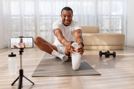 Photo for An enthusiastic African American man in athletic wear stretches on a yoga mat for a home workout, recording himself on a phone - Royalty Free Image
