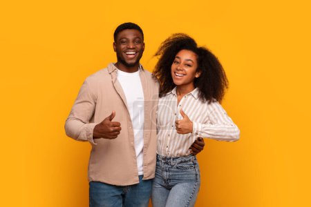 Téléchargez les photos : Souriant couple afro-américain debout côte à côte donnant pouces jusqu'à la caméra sur un fond jaune - en image libre de droit