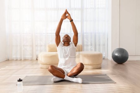 Téléchargez les photos : Tranquille homme afro-américain en tenue de sport confortable médite sur un tapis de yoga dans une pièce minimaliste - en image libre de droit