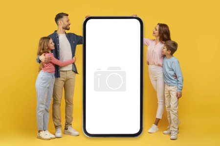 Foto de Familia alegre participar con un gran teléfono inteligente con blanco espacio de copia de pantalla en blanco maqueta en amarillo - Imagen libre de derechos