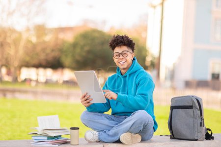 Photo for Cheerful young brazilian guy student wearing blue hoodie is using a digital tablet with a backpack and books around on a sunny day - Royalty Free Image