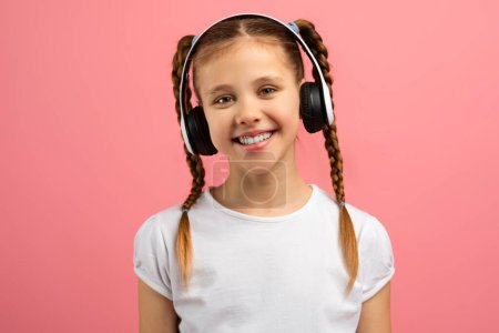 A delightful girl with a happy smile wearing headphones stands against a pink background, looking friendly