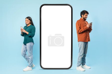Photo for Two cheerful young indian couple focused on their smartphones standing beside a large blank smartphone screen for mockups - Royalty Free Image