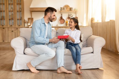 A father sits with his daughter enjoying a gift exchange moment, sharing a comfortable interaction on the sofa