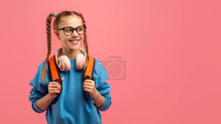 Joyful schoolgirl with glasses and headphones on a vibrant pink background, looking at copy blank space for ad