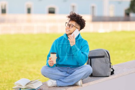 A young guy sits cross-legged on a ledge holding a coffee cup, talking on phone with books and a backpack nearby, portraying a casual outdoor study scene