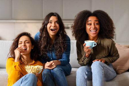 Three multiracial ladies friends enjoy a light-hearted moment, sharing laughter with a cup of coffee and popcorn bowl