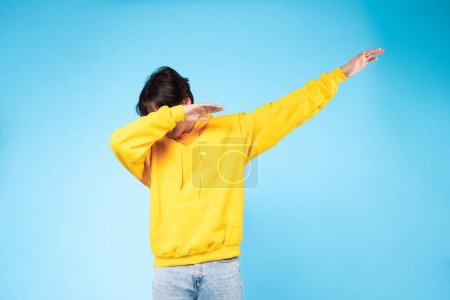 Téléchargez les photos : Une image dynamique capturant un mec à mi-teinte, avec un sweat à capuche jaune vif contrasté par un fond bleu solide, dégageant une ambiance cool et moderne - en image libre de droit