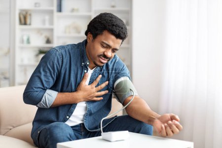 Ein erwachsener schwarzer Mann grimmig vor Schmerzen, seine Brust umklammert, während er seinen Blutdruck mit einem Home-Monitor überprüft, freier Raum