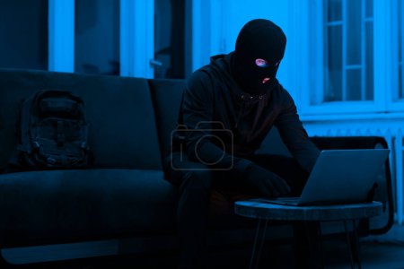 Téléchargez les photos : La lueur étrange de l'écran met en évidence le visage masqué d'un cybercriminel, donnant un aperçu des activités illicites en ligne la nuit - en image libre de droit