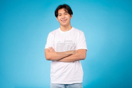Photo for A smiling handsome young asian guy confidently poses with crossed arms against a lively blue backdrop - Royalty Free Image