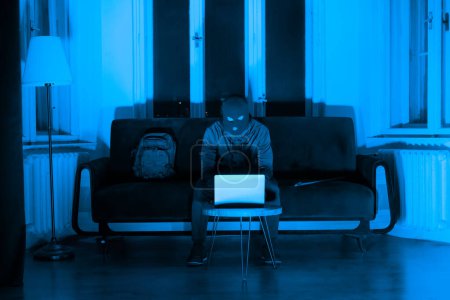 Un voleur à la recherche coûteuse avec un visage caché utilisant un ordinateur portable dans une pièce faiblement éclairée, suggérant une cyberactivité illicite