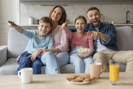 Una familia con dos hijos reacciona con entusiasmo a algo en el televisor mientras se sienta con aperitivos en un sofá