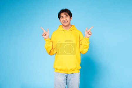 A joyful Asian teenage boy in a yellow hoodie posing with peace signs against a vivid blue studio background