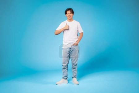 Photo for Smiling Asian teen in white tee and jeans gives a thumbs up, sharing a vibe of approval on a blue background - Royalty Free Image