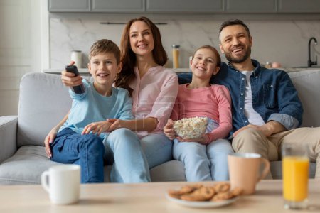 Téléchargez les photos : Une famille heureuse de quatre personnes s'assoit confortablement sur un canapé avec des collations, concentrées sur regarder la télévision ensemble - en image libre de droit