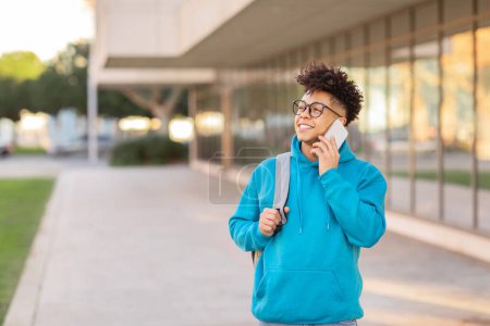 Téléchargez les photos : Un jeune homme noir marche sur un campus universitaire alors qu'il discute sur son téléphone pendant une journée ensoleillée - en image libre de droit