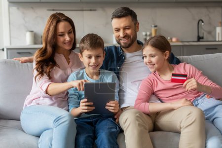 Vier Familienmitglieder sitzen auf einem Sofa und lächeln, während sie mit einem Tablet in ihrem Wohnzimmer interagieren, in der Hand eine Kreditkarte