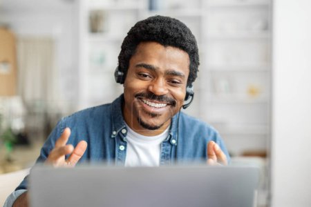 Téléchargez les photos : Amical homme noir avec casque à l'aide d'un ordinateur portable, engager une conversation joyeuse, travailler à la maison - en image libre de droit