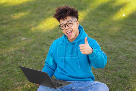 Foto de Alegre brasileño chico estudiante sentado en la hierba da pulgares hacia arriba mientras se utiliza un ordenador portátil, señalando una experiencia de estudio positiva al aire libre - Imagen libre de derechos