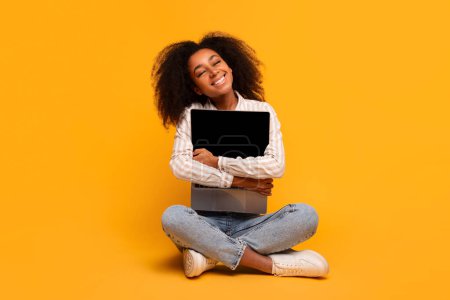 Foto de Una joven afroamericana con el pelo rizado sentada con las piernas cruzadas sobre un fondo amarillo vibrante, abrazando la computadora con la pantalla en blanco - Imagen libre de derechos