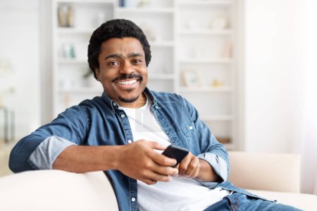 A cheerful black man sitting comfortably on a sofa while holding smartphone in a well-lit residential interior, smiling at camera