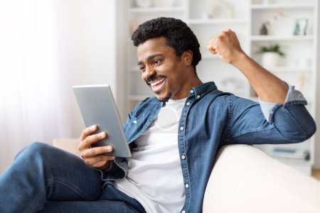 Téléchargez les photos : Un homme noir excité célébrant le succès tout en regardant sa tablette numérique, assis confortablement à l'intérieur à la maison - en image libre de droit