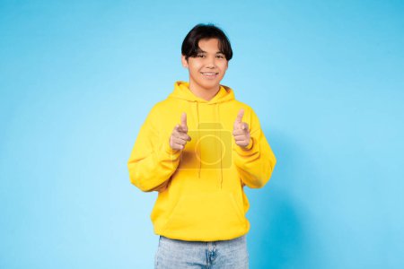 Foto de Feliz joven asiático chico en una sudadera con capucha amarilla apunta ambos dedos hacia la cámara con una sonrisa confiada en el fondo azul - Imagen libre de derechos