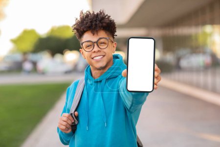 Glücklicher brasilianischer Typ Student in blauem Kapuzenpulli zeigt leeren Smartphone-Bildschirm in die Kamera, passend für eine Attrappe