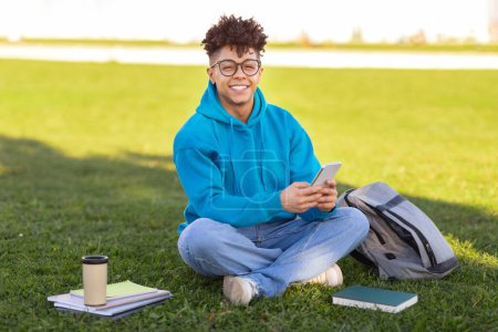 Téléchargez les photos : Young student in blue hoodie sits on grass with smartphone, textbooks, and coffee cup, exuding casual outdoor relaxation - en image libre de droit