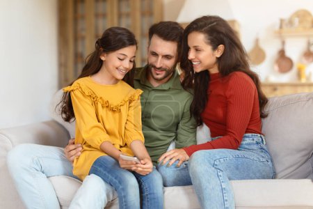 Téléchargez les photos : Joyeux moment de famille avec les parents et leur fille s'engageant avec un smartphone sur un canapé, une scène de vie domestique - en image libre de droit