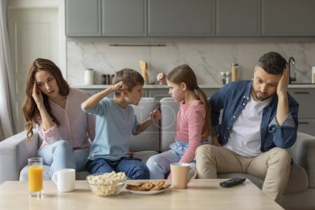Los hermanos niño y niña discuten sobre un juego mientras los padres se sientan sintiéndose estresados y cansados en una sala de estar