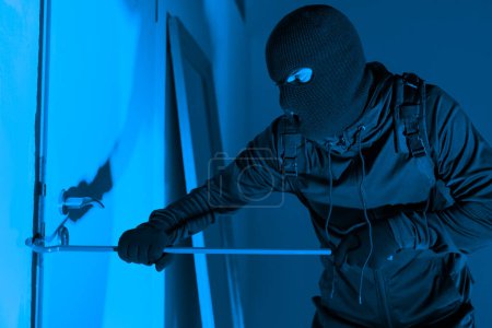 Téléchargez les photos : Image bleue d'un cambrioleur masqué en action utilisant un pied-de-biche pour ouvrir une fenêtre, suggérant un cambriolage nocturne - en image libre de droit