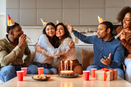 Foto de Amigos multirraciales con sombreros de fiesta se sientan alrededor de un pastel de cumpleaños con velas encendidas, compartiendo un momento de celebración - Imagen libre de derechos
