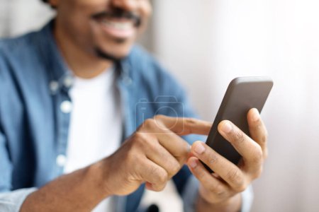Téléchargez les photos : Un homme noir joyeux interagit avec son smartphone, s'engageant avec un contenu qui lui apporte un sourire au visage, gros plan - en image libre de droit
