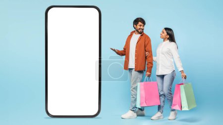 Photo for Hindu couple holds shopping bags beside an oversized blank screen smartphone signifying mobile shopping against a blue backdrop - Royalty Free Image