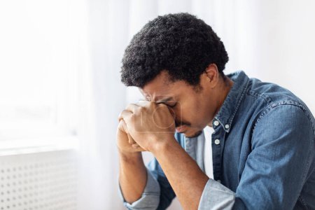 Foto de Un joven negro está sentado enterrando la cara en su mano, exudando una sensación de frustración o angustia en casa. - Imagen libre de derechos