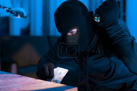 A thought-provoking image of a masked burglar counting possibly stolen money in a dimly lit scene