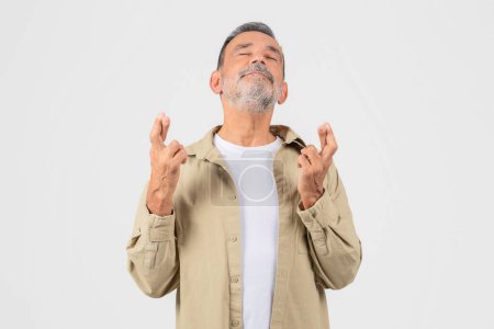 Photo for An elderly man stands with hopeful expression crossing his fingers wishing for good fortune or a positive outcome in an uncertain situation - Royalty Free Image
