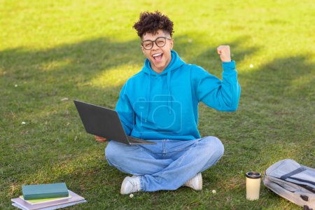 A joyful brazilian guy student sitting on grass with a laptop, celebrating success, notebooks and a coffee cup alongside