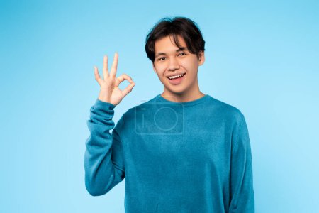 gai les jeunes asiatique guy avec un rayonnant sourire montrant un ok main signe dans un bleu studio toile de fond